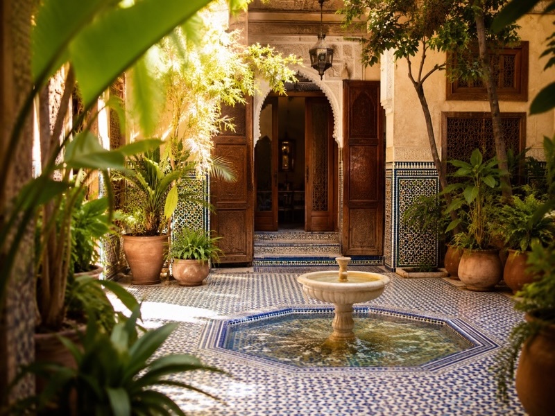 Sonnendurchfluteter Innenhof eines traditionellen Riads in Marrakesch mit blauen Kacheln, grünen Pflanzen und einem zentralen Brunnen